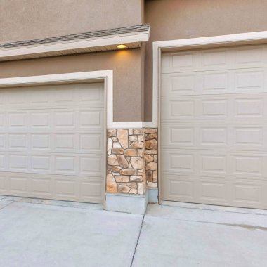Square Three-car garage exterior with light brown sidings. There are two sectional garage doors with concrete driveway and a siding made of concrete and stone veneer siding.