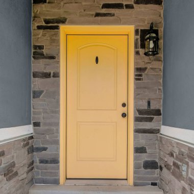 Square Yellow front door with doorbell and porch lamp on the side. Entrance exterior of a house with railings at the front and gray concrete and stone veneer siding.