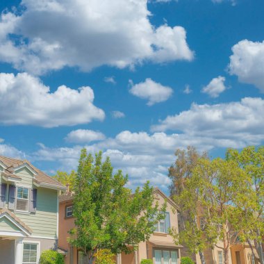 Square White puffy clouds Row of traditional houses at Ladera Ranch in Southern California. There are trees at the front of the houses against the clear sky background.