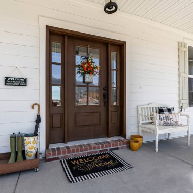 Square Porch of a house with bench and wooden rack with boots and umbrella. There is a white vinyl wood siding, window with shutter design and front door with sidelights and hanging flowers