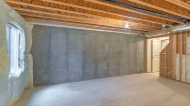 Panorama Unfinished basement with a plastic vapor barrier on the wall. Empty basement interior with wooden frameworks and insulated wall on the left and a window on the right.