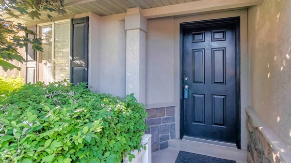 Panorama Front porch of a house with shrubs and trees at the front. There is a white railing near the shrubs and a black front door with lockbox and black doormat at the concrete flooring.