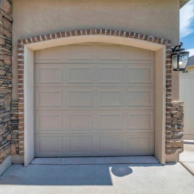 Square White puffy clouds Beige sectional garage door with arched door opening. Garage exterior with stone veneer siding and bricks doorframe against the vinyl fence at the back.