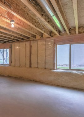 Vertical Unfinished basement with plastic vapor barrier and windows. Interior of a basement with wooden ceiling beams with metal pipe and light bulbs.