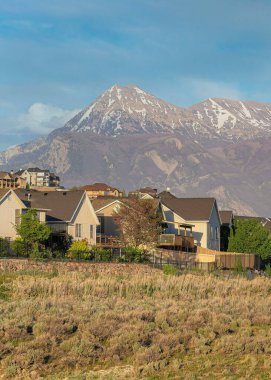 Vertical Whispy white clouds Vast of shrubs and grass with dirt road at the front of residential area. There are colorful residential houses against the mountain range and sky at the background.