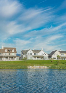Vertical Whispy white clouds Oquirrh Lake waterfront at Daybreak, Utah residential area. Reflective water of the lake against the large home facades against the mountains and clear blue sky.
