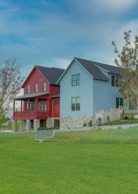 Vertical Whispy white clouds Three large two-storey houses with colorful wood sidings at Daybreak, Utah. There is a lawn with benches at the front of the white picket fences of the red