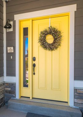 Vertical Yellow front door with wreath and sidelight at the entrance of a house in Utah. Home exterior with gray wood vinyl and stone veneer sidings and a porch with wall lamp near the window.