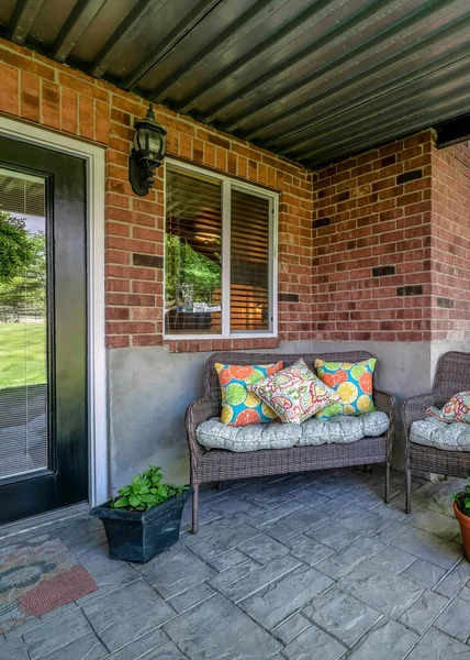 Vertical Woven couch and armchairs on an outdoor patio under the deck of a house with bricks. There is a glass front door on the right side near the windows and a view of a stairs on the left.