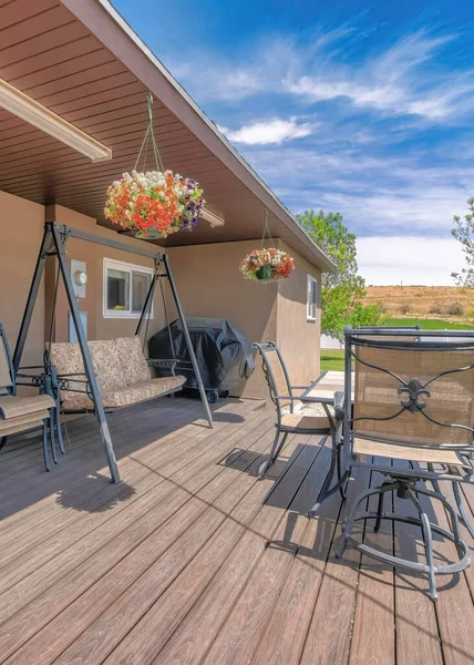 Vertical Outdoor patio with covered barbecue grill, wooden flooring and lounge chairs. There are stacked of chairs beside the hanging couch near the sliding glass door and flowers hanging