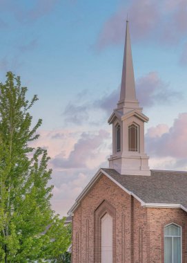 Vertical Puffy clouds at sunset Top exterior of a church in Utah with trees outdoors. Church exterior with three tone bricks siding and a steeple tower at the front.