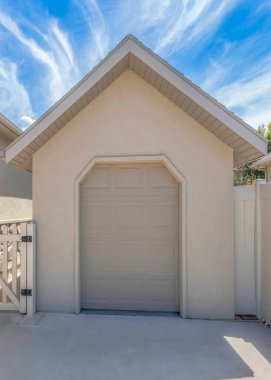 Vertical Whispy white clouds Tool shed with clipped edge entrance and sectional door. There is a picket gate on the left near the house with stone veneer sidings and a vinyl fence on the right.
