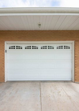 Vertical White sectional garage door with glass panels at the top. Exterior of a house with brown bricks siding and a concrete driveway at the front of the garage.
