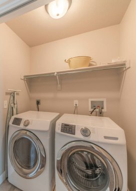 Vertical Reached in laundry with white bi-fold doors and laundry units below the shelf. Interior of a house with laundry room near the stairs and room with window and carpeted flooring.
