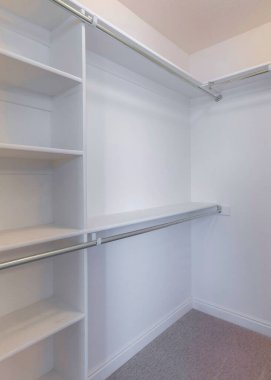 Vertical Small white walk-in closet with brown carpet flooring. There is a shelving unit with metal rods on the left and hooks on the right.