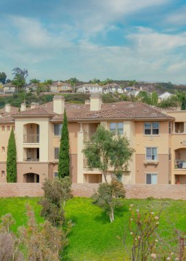 Vertical Whispy white clouds Fenced complex apartment buildings with balconies at San Diego, California. There is a lawn and trees near the stone fence and a view of an uphill road