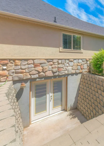 Vertical Whispy white clouds Basement entrance with glass panels on the double doors. There is a stairs near the stack of concrete blocks near the lawn and a view of bushes and trees