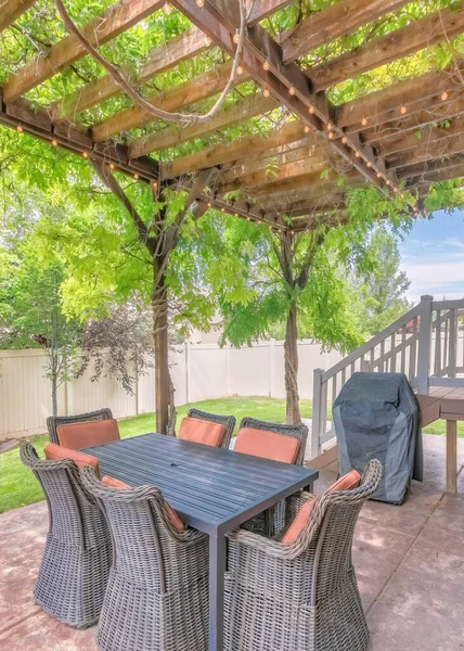Vertical Outdoor patio with covered barbecue grill under a pergola roof with vines. There are woven chairs tucked into table near the wooden deck on the side and a view of a vinyl fence and lawn.