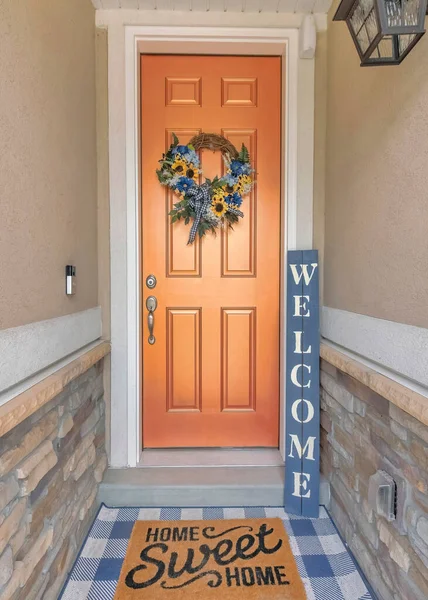 Vertical Bronze front door with wreath, welcome signage and doormat. Entrance of a house in the middle of the walls with stone veneer siding at the bottom.