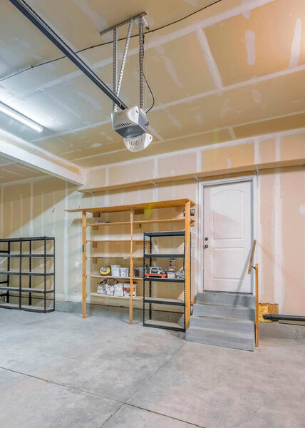 Vertical Large unfinished garage interior with wall patch marks. There are two automatic door operators on the ceiling and storage racks and shelving