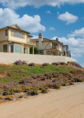 Vertical White puffy clouds Dirt road outside the fenced beach houses at Carlsbad, California. There are large houses on the left with white fence near the bushes and a view of the sea at the right.