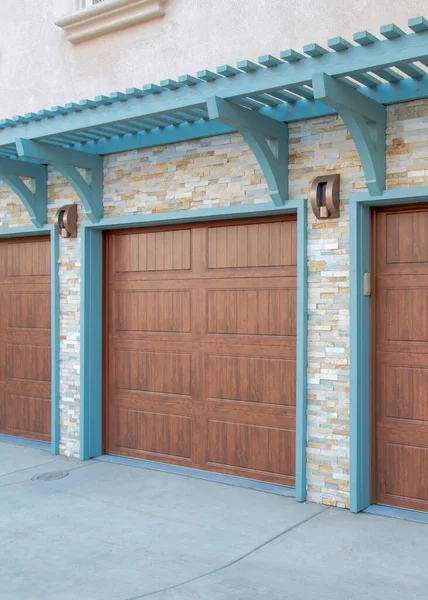 Vertical Garage exterior with wall-mounted light blue pergola and doorframe. There are three wooden sectional doors and stone veneer wall sidings.