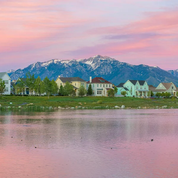 Square Reflective Oquirrh Lake waterfront Daybreak, Utah. Panoramic view of the lake at the front of the houses and fields against the mountain range and sky with pink hue.
