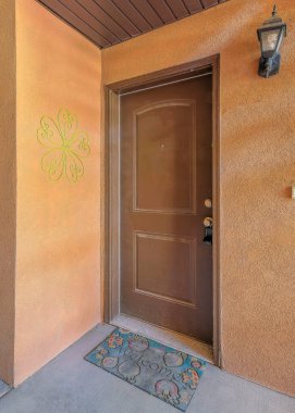 Duvar süslemesi, duvar lambası ve paspası olan dikey kahverengi bir ön kapı. Evin girişi güvertenin altında, kahverengi tavanlı ve her iki tarafında iki merdiven manzaralı..