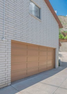 Dikey beyaz kabarık bulutlar garajın yanındaki kıvrımlı garaj yolu kaygan pencereli ve beyaz desenli tuğlalı duvar. Komşu ev ve dağ evinin tepesinde kahverengi vinil çitli beton bir duvar var..