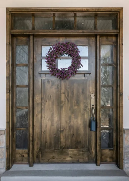 Vertical Large dark wood front door with two side panels and transom window. Home entrance with lavender wreath on the front door and an exterior with gray vinyl wood and stone veneer sidings.