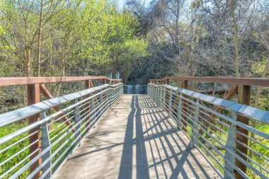 Boardwalk over the wild plants on the side with trees at Austin, Texas. Walkway with concrete path and metal barriers leading to a two trash cans at the back.