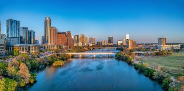 Colorado River near the district urban area of Austin, Texas. There are bridges over the river and a view of skyscraper buildings on the left and large field at the right side against the sunset sky.