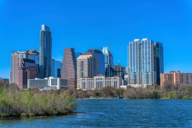 Austin, Texas cityscape and view of Colorado River at the front. There are trees at the shore of the river near the buildings against the clear blue sky at the back.