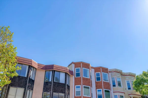 Rowhouses with bay windows against the clear sky in San Francisco, CA. There are two houses from the left with black left and brown center wood sidings beside the beige house on the right.