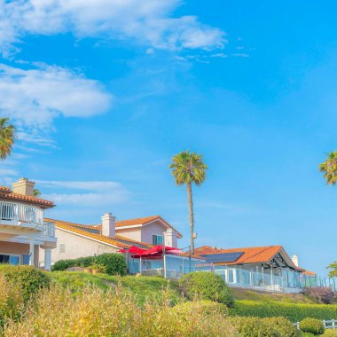 Kare beyaz kabarcık bulutlar Güney Kaliforniya 'da büyük evler ve palmiye ağaçları. Evin sol tarafında terası olan bir ev var. Sağ tarafında güneş panelleri ve cam çitler var..