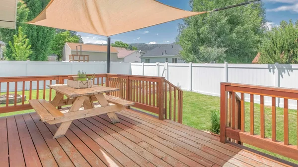 Panorama Whispy white clouds Wooden deck with a sunshade over the table with bench seats. Deck at the backyard of a house with a view of a lawn and white vinyl fence against the trees and neighborhood.