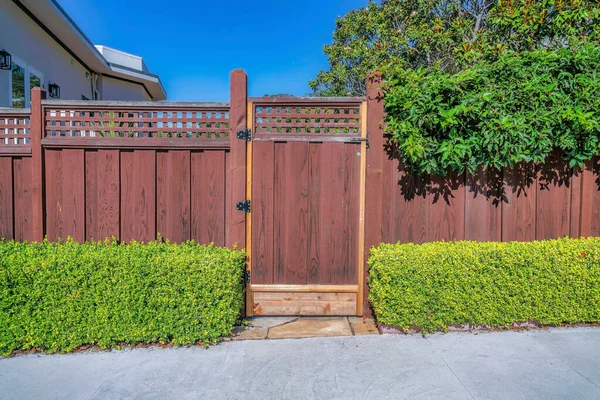 Closed gate at the entrance of a home with blue sky background on a sunny day. Brown wooden gate and fence with metal lattice with landscaped plants at the entryway of house.