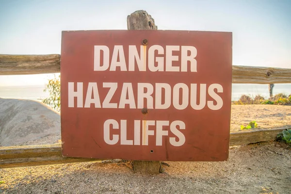 Danger Hazardous Cliffs sign by the beach at Del Mar Southern California. Close up view of a safety warning sign with sandy shore, sea and sky scenery background at sunset.