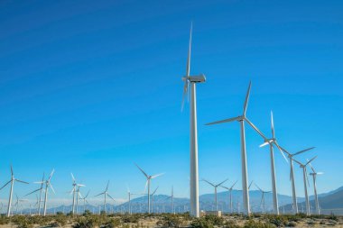 Dağları ve açık mavi arkaplanı olan yel değirmenlerinin güneşli manzarası. Yenilenebilir alternatif enerji kaynağı olan rüzgar pompaları alanı.