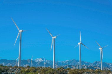 Dağlara karşı üç keskin bıçaklı ve mavi arka planlı yel değirmenleri. Kuru kayalıklarda çalılarla güneşli ve güneşli bir gün..