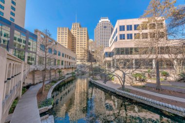 San Antonio Teksas 'taki nehir yürüyüşünün güzel manzarası binalara karşı. Turistlerin yürüyebileceği ve manzaranın keyfini çıkarabilecekleri kaldırımlar mavi gökyüzünü yansıtan kanalın etrafını saran.