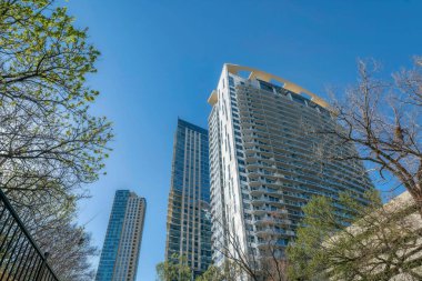 Austin Texas şehir merkezi, dış görünüşü modern binalar ve ağaçlar. Yükselen gökdelenlerin ön cephesinde mavi gökyüzünü yansıtan cam pencereler vardır..