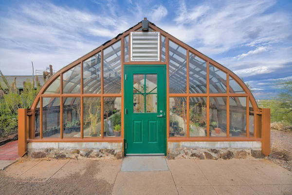 Gothic arch greenhouse with glass panel and green door below the stainless ventilation at Tucson, AZ. Front exterior of a greenhouse with painted woodframes against the sky background.