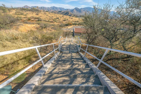Concrete outdoor stairs with metal railings on a mountain with wild plants and trees- Tucson, AZ. View of a staircase from above with a view of a field and mountains at the back.