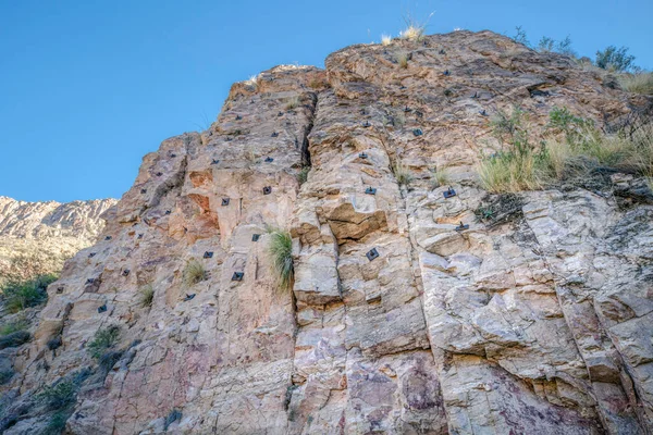 Rocky mountain cliff with rock climbing anchor bolts at Sabino Canyon State Park- Tucson, AZ. View of a steep rock wall cliff with grasses in between the cracks against the sky.