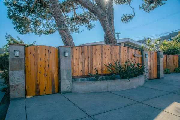 La Jolla, California- Wooden gates and walls with tiled columns. Front of a property with planters at the front of the wooden wall fence against the two large trees at the background.