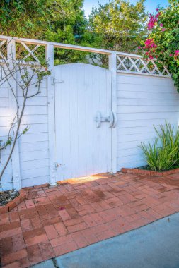 La Jolla, California. Beyaz tahta kapı ve çit boyalı. Kaldırımda kırmızı tuğlalar, yan tarafında bitkiler ve arka tarafında ağaçlar olan bir giriş..