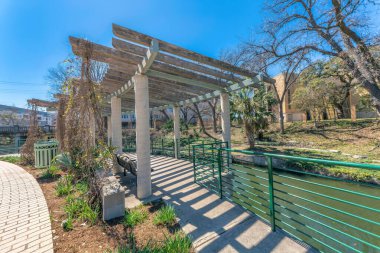 San Antonio, Teksas 'ın aşağısında nehir kenarındaki beton yolda ağaçlarla yürüyoruz. Yol ve nehir arasında kurumuş asmalar ve korkuluklar olan bir pergola çatısı var..