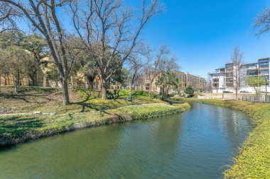 San Antonio, Teksas şehir merkezi. Kenarlarında bitkiler, binalar ve gökyüzü manzarası olan nehir yürüyüşü. Nehrin karşısında binalara karşı yapraksız ağaçlar ve arkada mavi gökyüzü var..