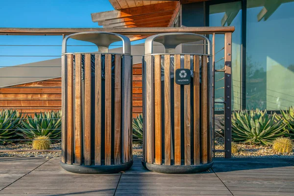 La Jolla, California- Two trash cans with wood planks. Wooden trash storages with recycle bin on the right against the cable railings and succulent plants at the front of a building with glass wall.